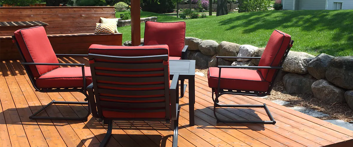 Cedar Decking Installation in Blue Bell Outdoor cedar deck with red cushioned chairs and a table, surrounded by lush green lawn and rock landscaping.