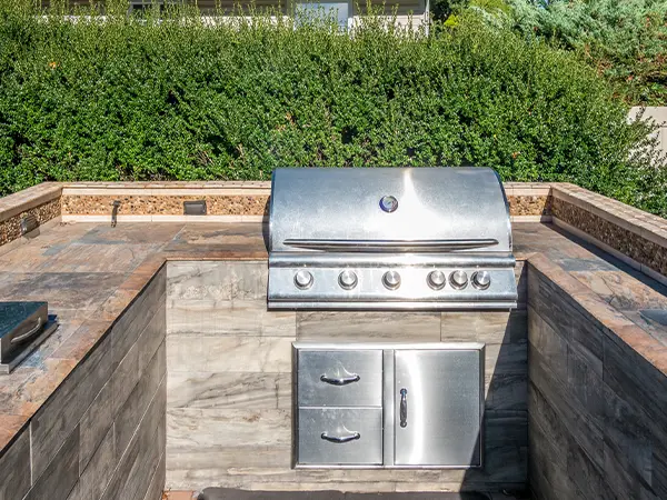 Outdoor stainless steel grill built into a stone countertop with a green hedge background