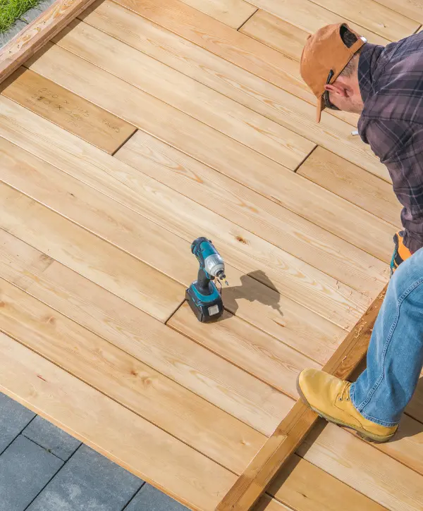 Man in workwear kneeling on a partially built wooden deck, with a cordless drill placed nearby