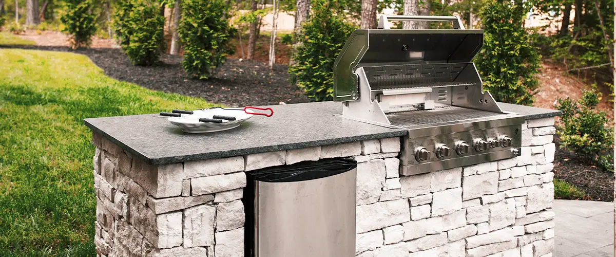 Outdoor grill with stone base and granite countertop on a patio, surrounded by a landscaped backyard