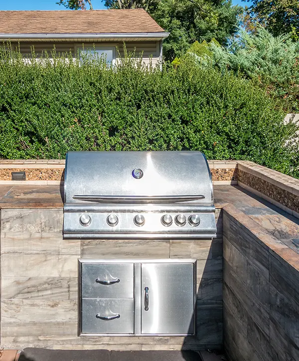 Outdoor stainless steel grill built into a stone countertop with a green hedge background