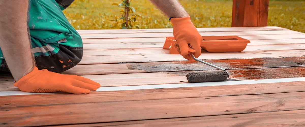 Person applying stain to a wooden deck using a roller, wearing orange gloves