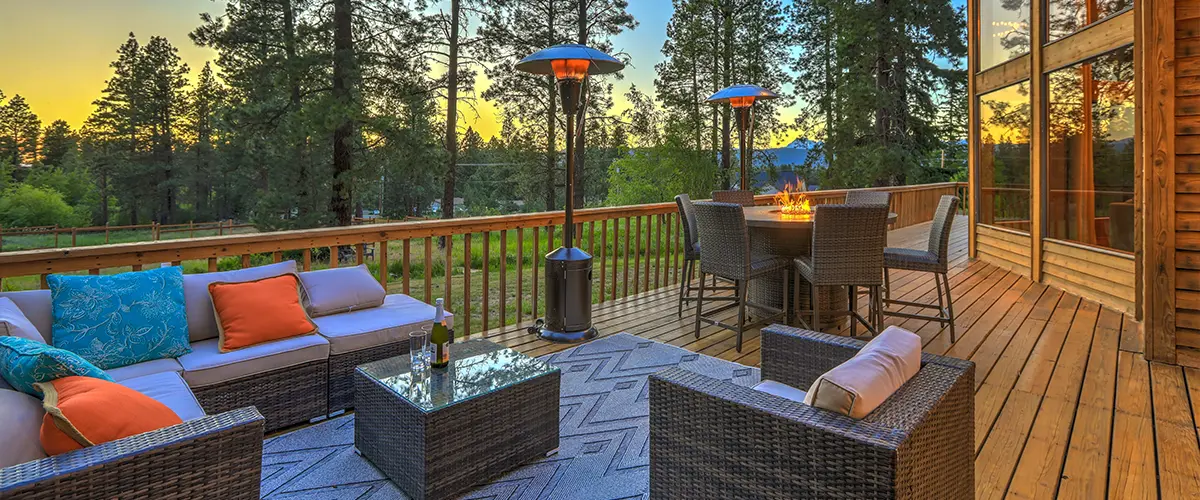 Cozy outdoor deck with wicker furniture, fire pit, patio heaters, and forest view at sunset - modern backyard entertaining area.