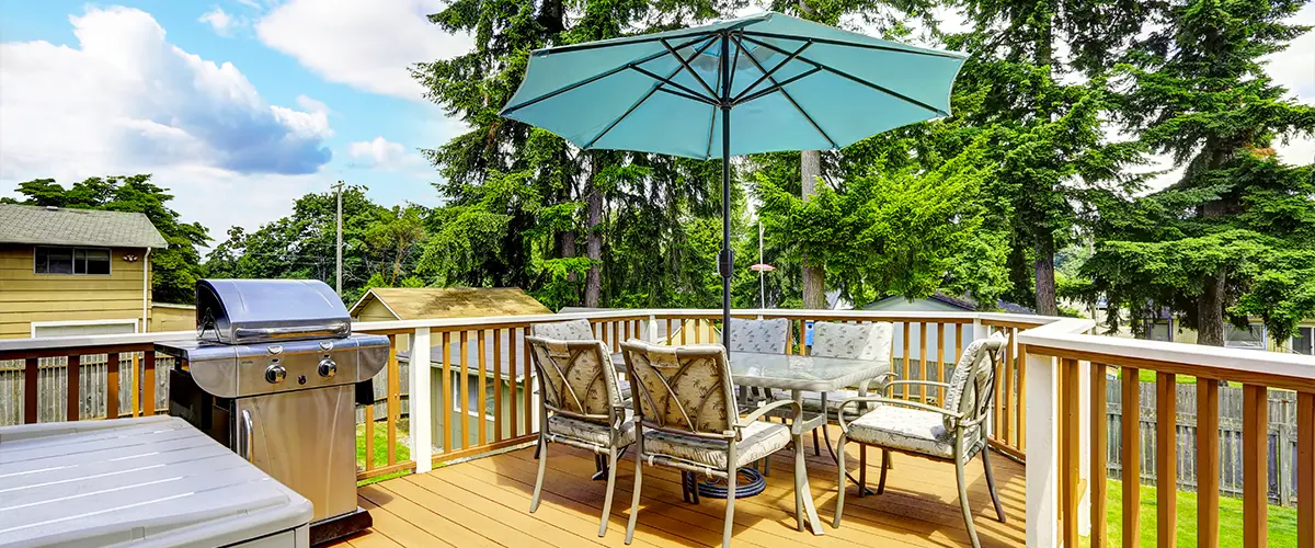 Outdoor deck with patio furniture, teal umbrella, and stainless steel BBQ grill surrounded by trees and a wooden railing.