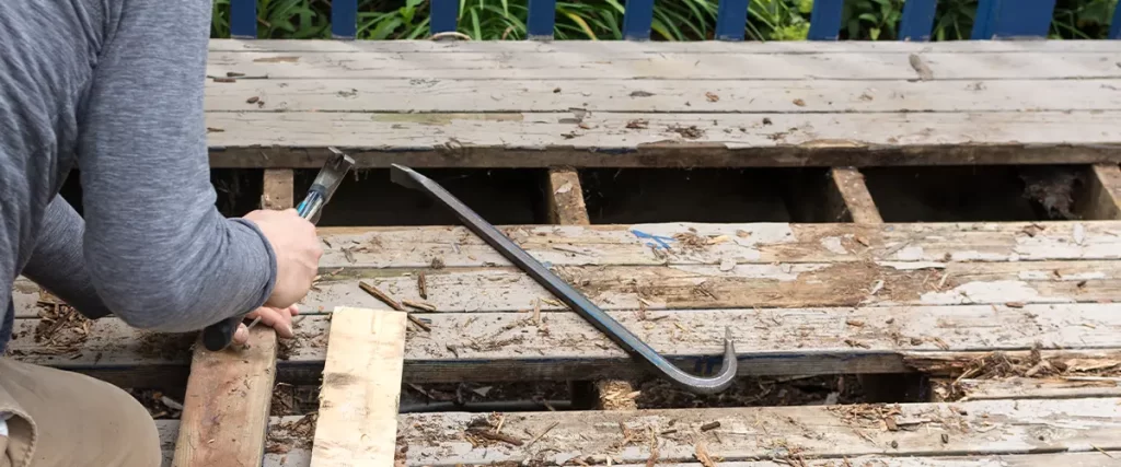 Worker repairing a rotted wooden deck, using tools to replace damaged boards for improved deck safety and durability.