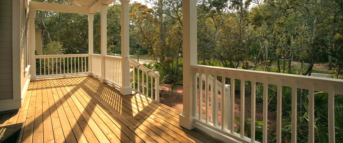Charming covered front porch with white railings and wooden deck overlooking lush trees and landscaped front yard.