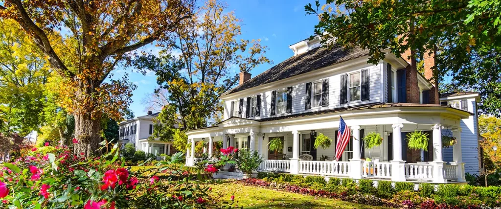 Elegant colonial-style home with white wraparound porch, black shutters, American flag, and blooming front garden