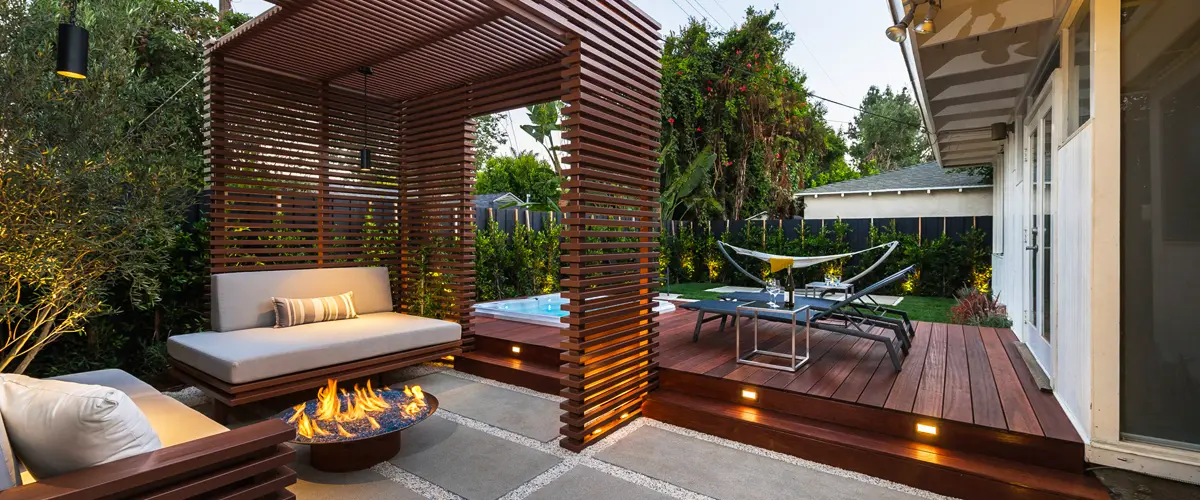 Modern backyard lounge with fire pit, wood slat pergola, and raised deck featuring hot tub and lounge chairs.